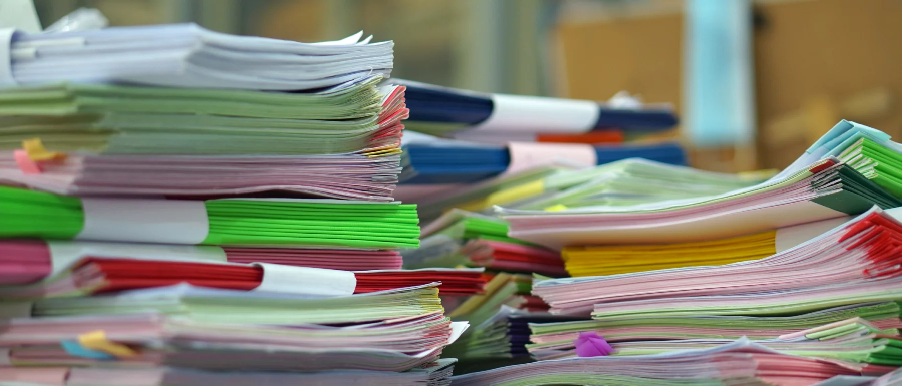 A close-up image of multiple stacks of documents and files arranged in piles on a surface. The documents are held together with colourful plastic binders and clips.