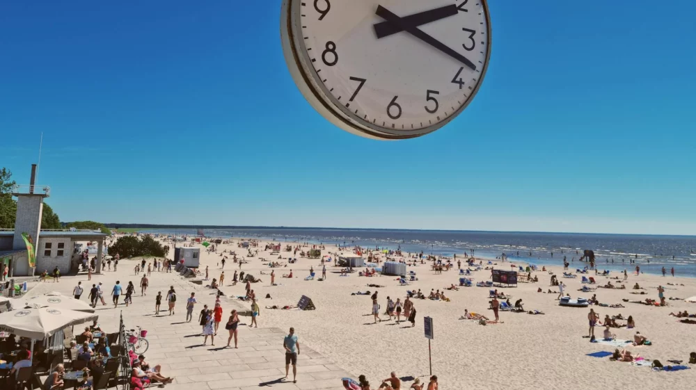 A seaside beach with a lot of people and a clear sky above. The photo is taken from a building that features a large analog clock, which is displayed on the photo above the people.