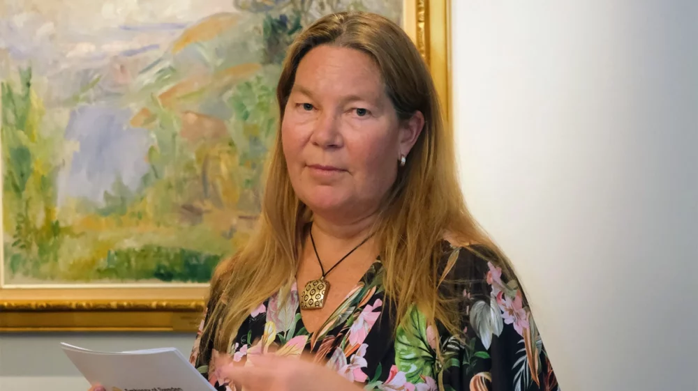 Annika Markovic holding papers, standing in front of a framed painting.