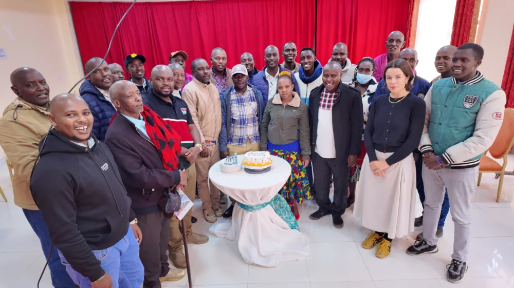 Group photo of around twenty people standing indoors around a table with a cake.