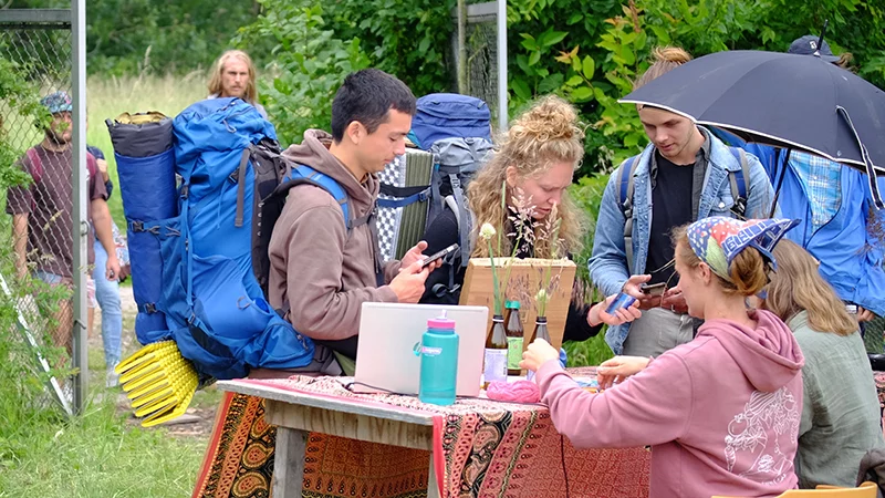 Young people with backpacks registering at an outdoor table covered in a colourful cloth, using phones and filling out forms, with tents and trees in the background