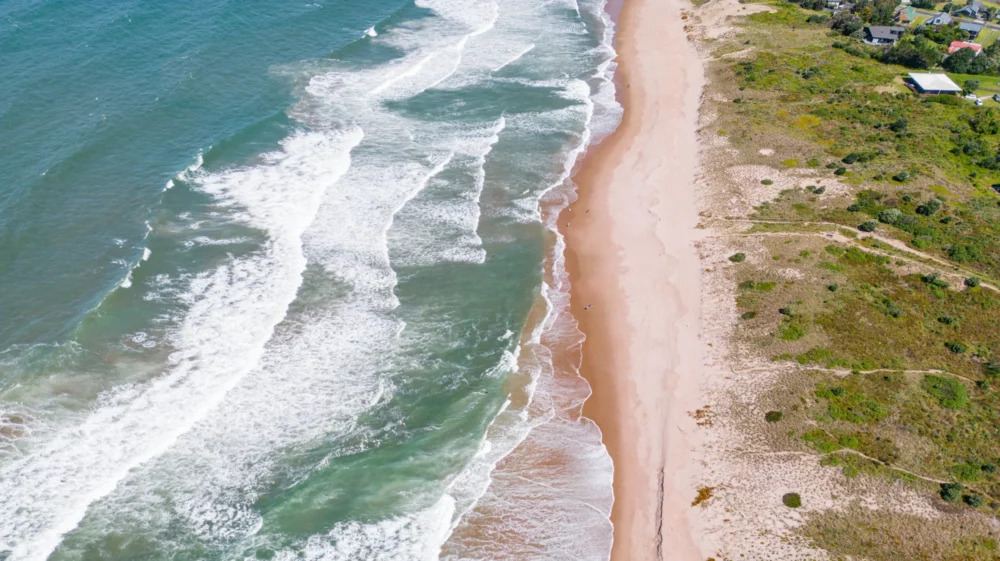 Aerial view of ocean waves breaking along a sandy beach bordered by green dunes and scattered houses.