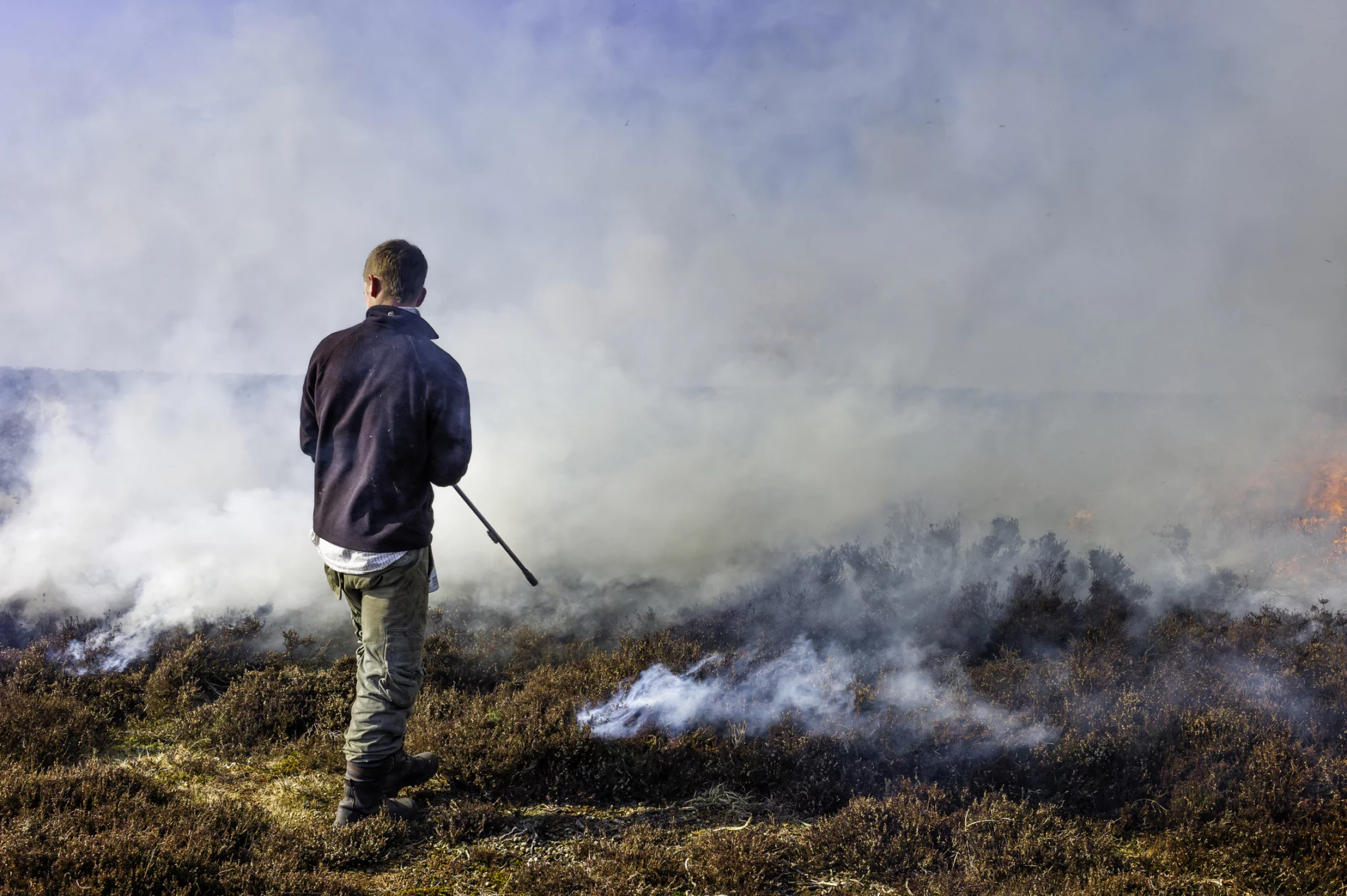 A man stands on heather moorland with his back to the camera. Directly in front of him, parts of the ground billow smoke as he administers a controlled burn of the heather for land management.