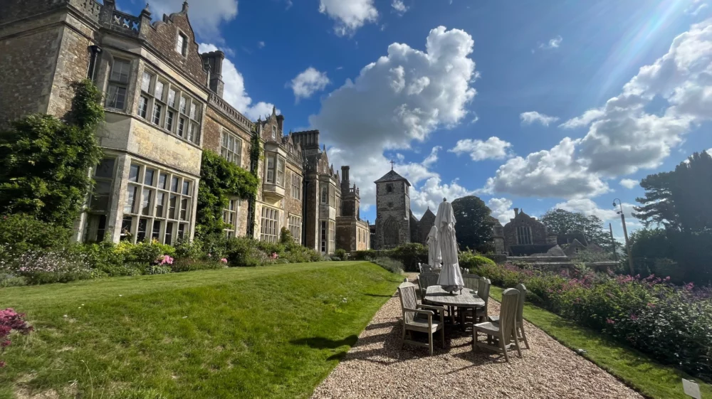 Blue sky with white piled clouds above a formal stone residence with a path and table, chairs and closed umbrella surrounded by green grass.