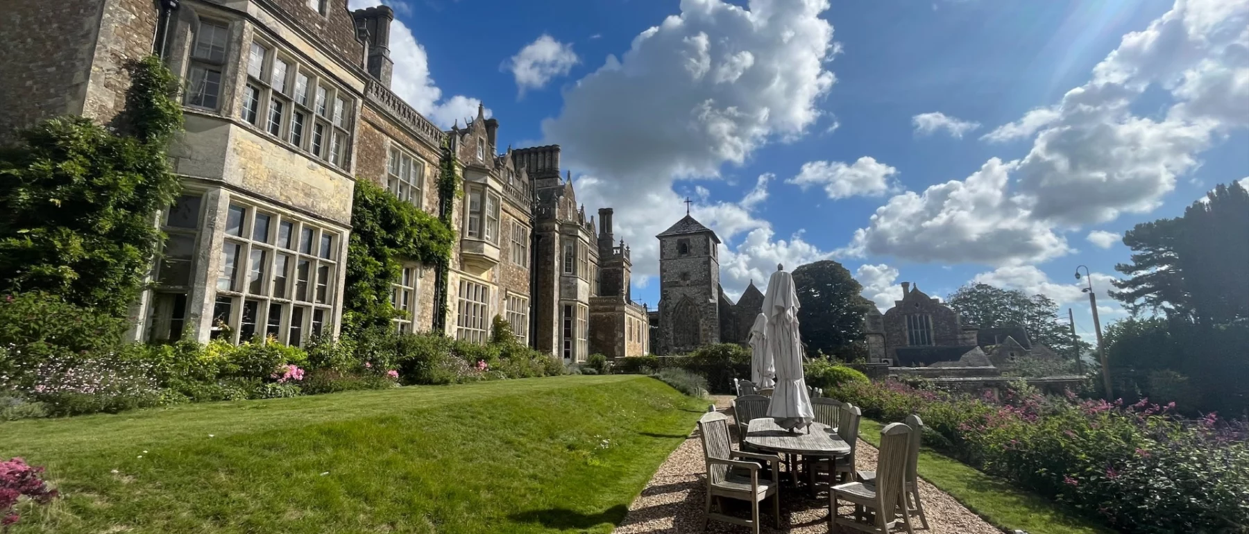 Blue sky with white piled clouds above a formal stone residence with a path and table, chairs and closed umbrella surrounded by green grass.