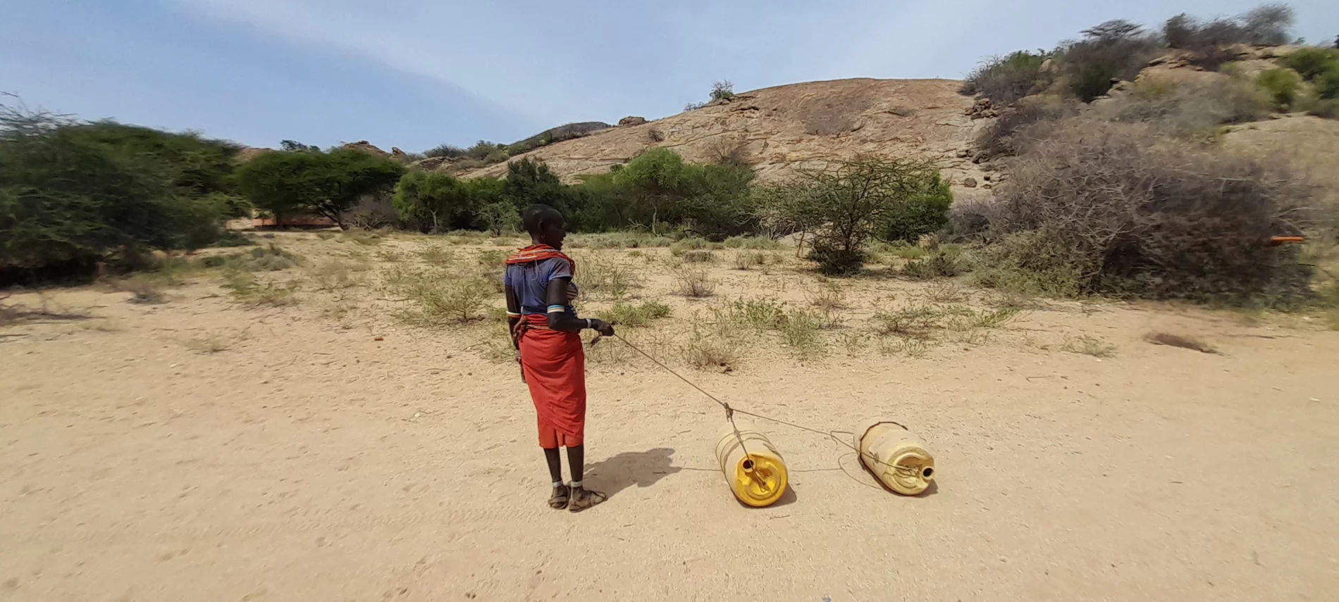 A person in red with back to camera looks to the right where two yellow plastic cylinders lie on sandy ground, with scrub bushes in the background.
