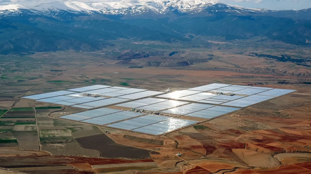 Shimmering squares tile the brown and green land, with blue snow-capped mountains and clouds on the horizon under a sunny blue sky.