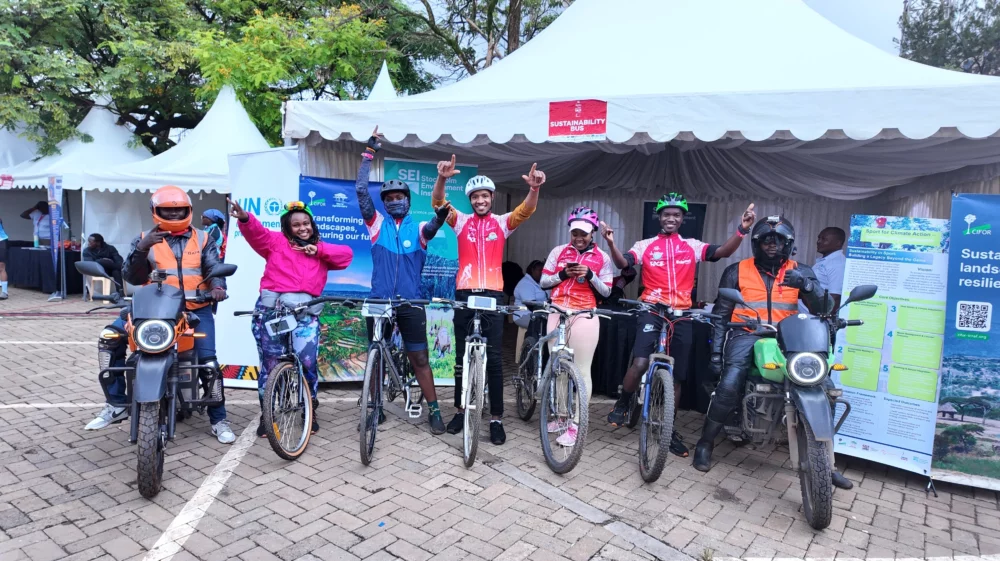 Group of cyclists and electric motorbike riders smiling and raising their hands in front of the SEI “Sustainability Bus” tent at the Grand Nairobi Bike Race.