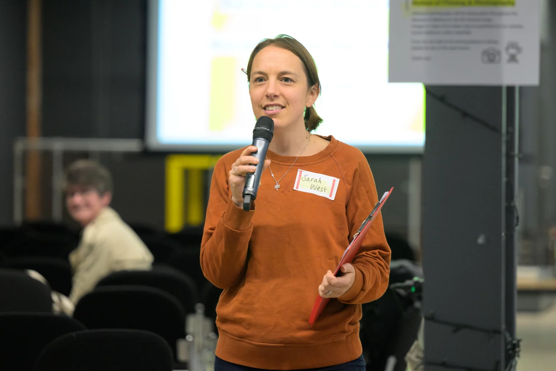 A woman holding a microphone and a red clipboard speaks at an indoor event. She is wearing an orange jumper and has a name tag that reads “Sarah West.” The background shows a seated audience member and a blurred presentation screen.