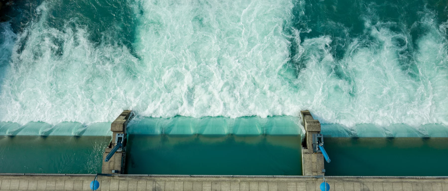 Overhead view of a dam where the water barrage flows into a turquoise body of water