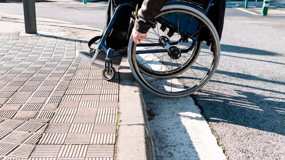 Half-body details of a man sitting in his wheelchair climbing up a curb that is not wheelchair accessible.