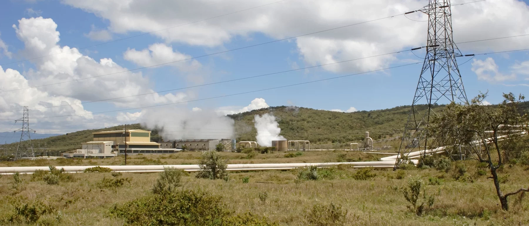 A large industrial complex in yellow and tan sprawls on a hill of dry grass and scrub, with white plumes of steam and electricity lines and two electrical towers above under a blue sky with white clouds.