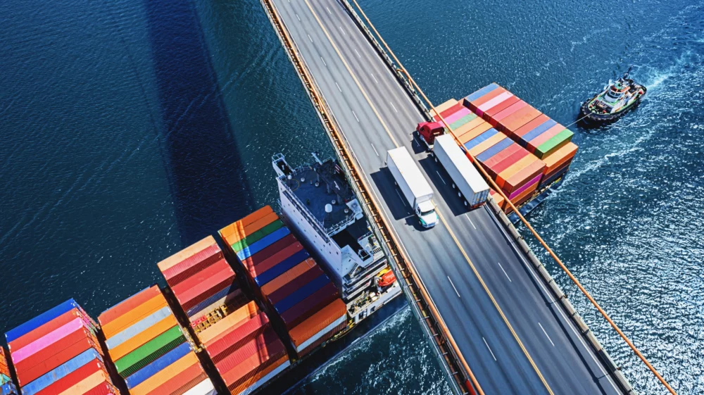 Aerial drone view of a container ship navigating beneath two trucks crossing a suspension bridge.