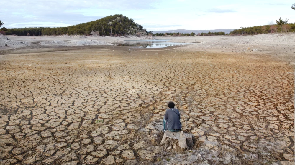 A person sits with back to the camera in an expanse of dried lake bottom, with cracked mud into the distance where a small pond sits near what was an island at high water.