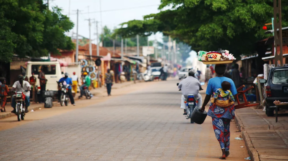 Street scene in an African town with people walking and riding motorcycles, and a woman carrying goods on her head and a baby on her back.