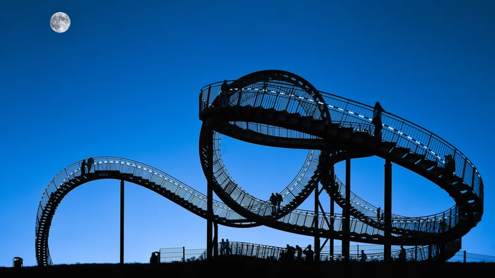 Silhouette of the walkable roller-coaster sculpture ‘Tiger & Turtle – Magic Mountain’ in Duisburg, Germany, with people on its looping ramps under a twilight sky and a visible moon.