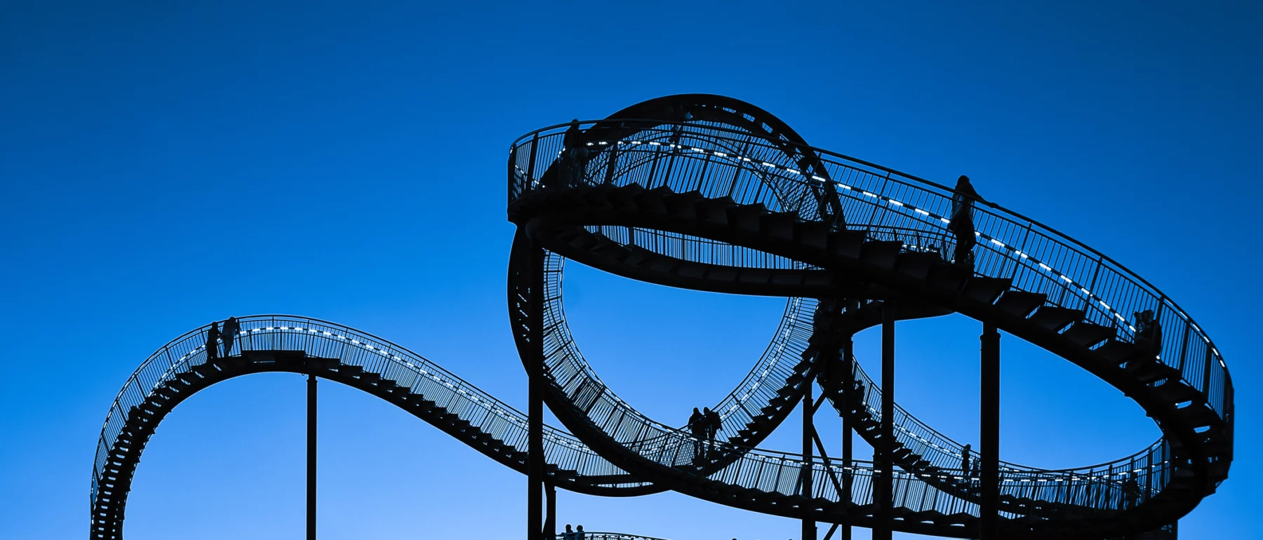 Silhouette of the walkable roller-coaster sculpture ‘Tiger & Turtle – Magic Mountain’ in Duisburg, Germany, with people on its looping ramps under a twilight sky and a visible moon.