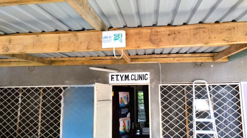 A door with a sign above it that says FTYM clinic, with an air monitor above the door with logos for SEI and UNEP, with grilled windows and wooden beams under the overhanging roof and a ladder.