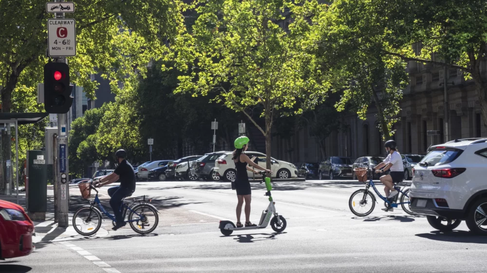 Cyclists and an e-scooter rider waiting at a city intersection on a sunny day.