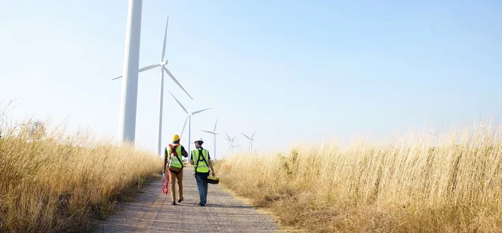 This image depicts two engineers walking along a path beside a farm of wind turbines in Thailand.