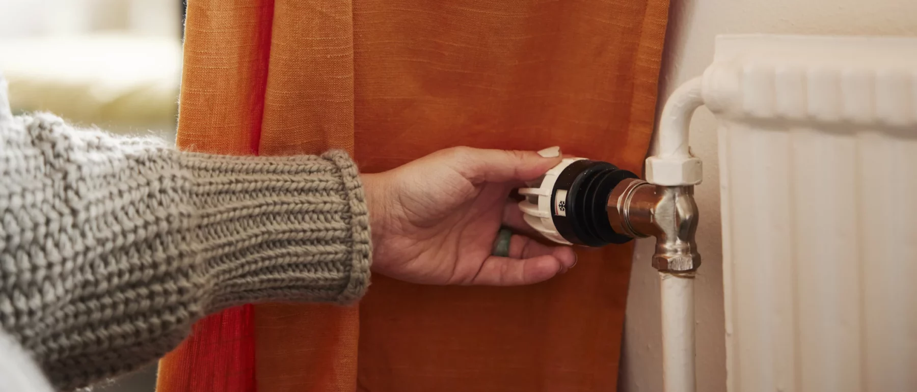 Hand in a knitted sleeve adjusting a radiator thermostat behind an orange curtain.