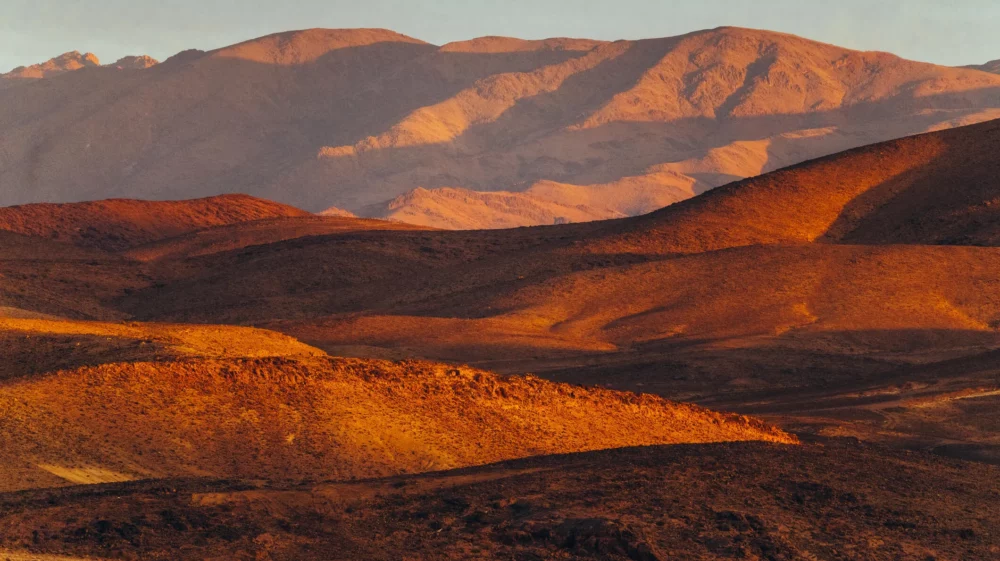 Sunlit desert hills with rugged mountains in the background at sunrise or sunset.