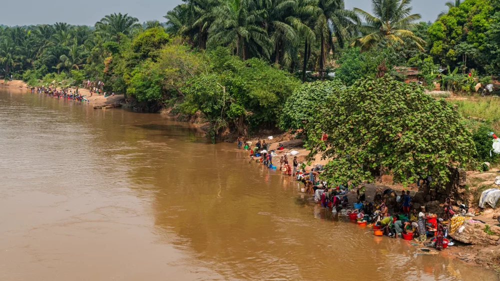 People gathered along a muddy riverside washing clothes beside dense tropical vegetation.