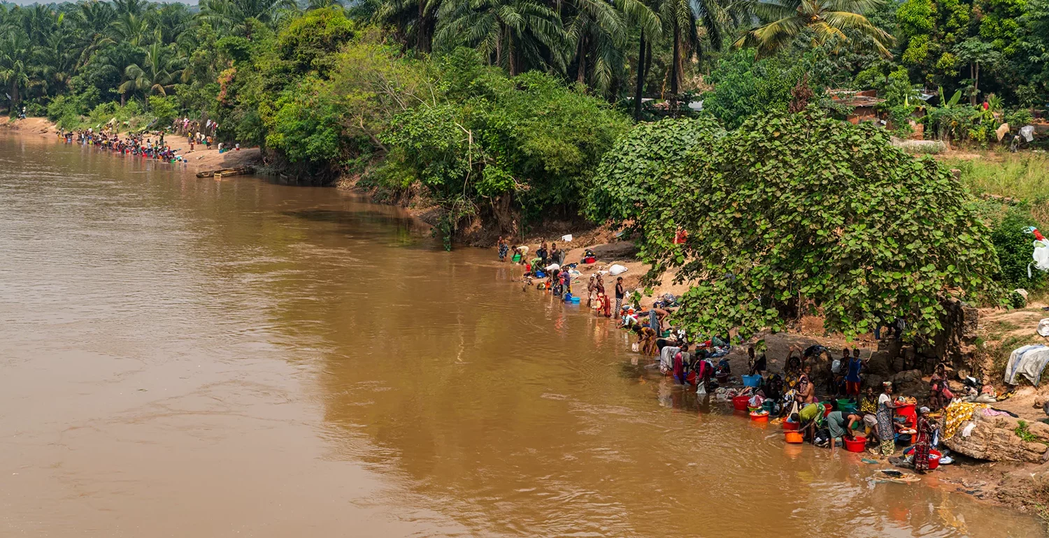 People gathered along a muddy riverside washing clothes beside dense tropical vegetation.