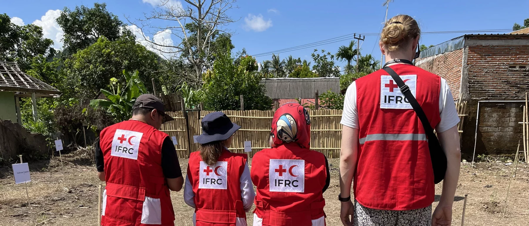 Four people wearing red IFRC vests stand with their backs to the camera, looking at a dirt field.