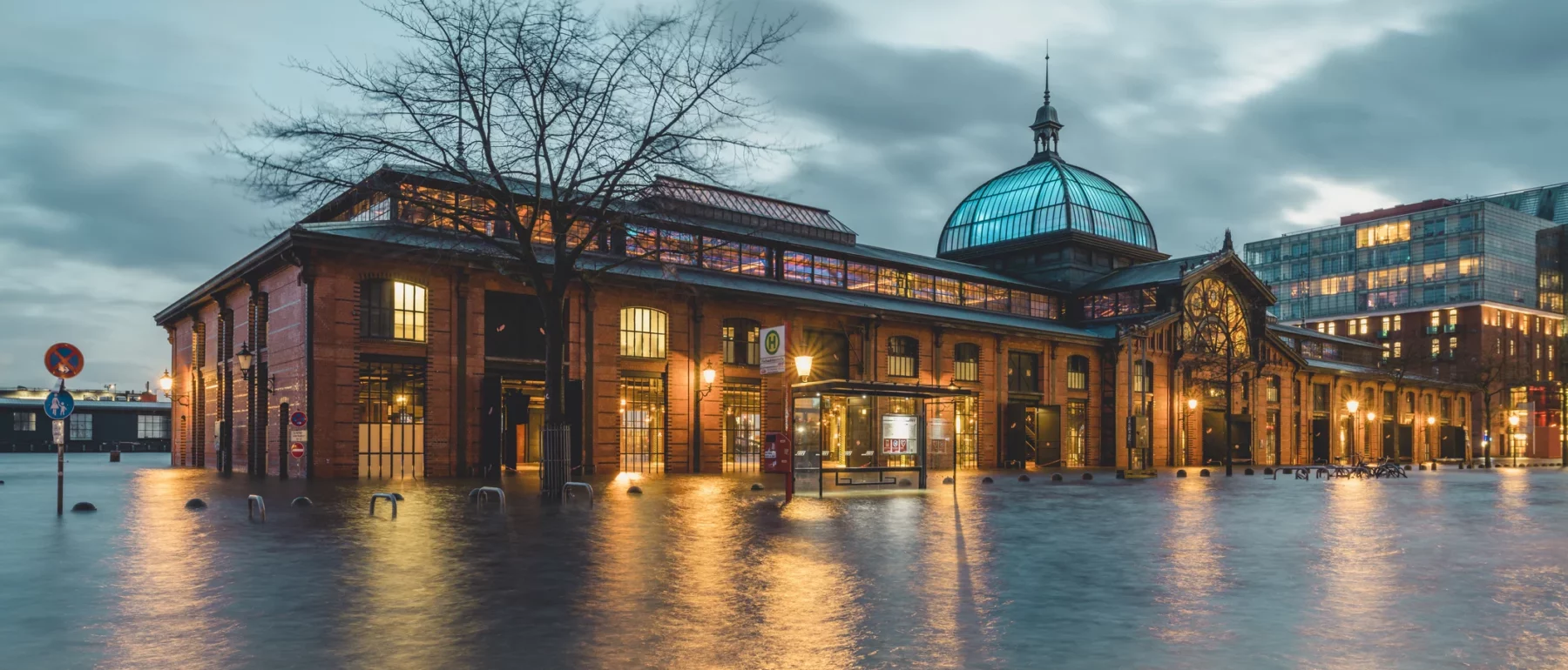 A long brick building with a glass cupola on the roof sits in floodwaters halfway up the lower floor windows, with lights spilling out and mirrored on the surface, a skeletal tree, gray cloudy sky, and city skyline in the background.