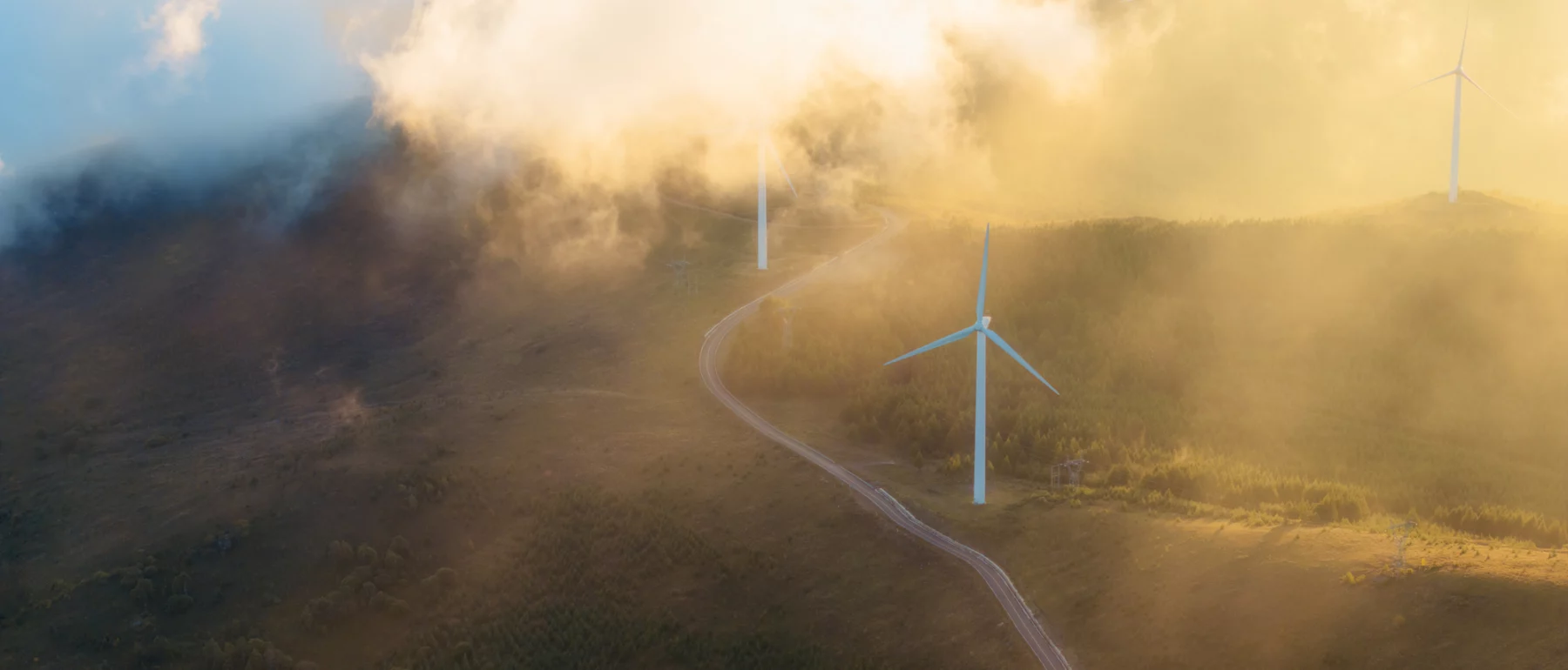 Wind turbines stand along a winding road in a hilly landscape, partially shrouded in low clouds and golden light, illustrating renewable energy development in a natural setting.