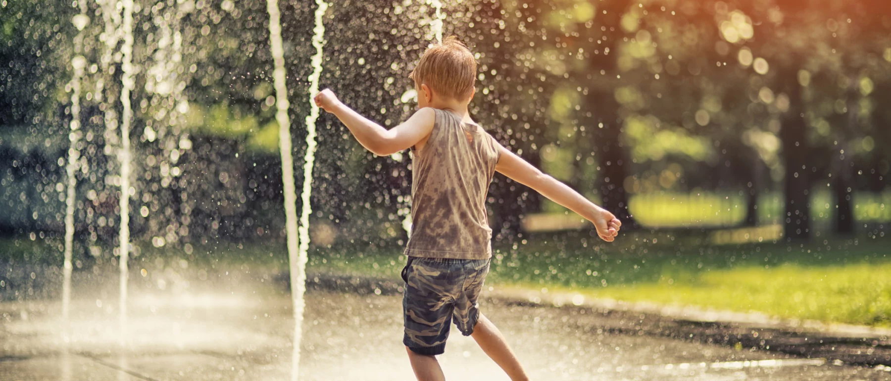 Hot summer day in the city. Little boy is having fun playing in the park fountain. He is running barefeet getting wet and splashing.