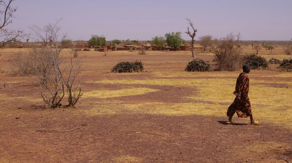 A man on the right side of the image walks across a dry yellow and brown landscape, with occasional brown and green scrub brush and green trees at the horizon that stand among a cluster of clay brick houses, some square with flat roofs, some cylindrical with conical metal roofs.