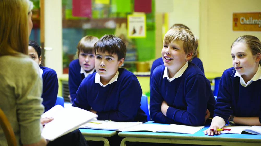 A group of primary school children in blue school jumpers sitting at desks in a classroom and facing a teacher.
