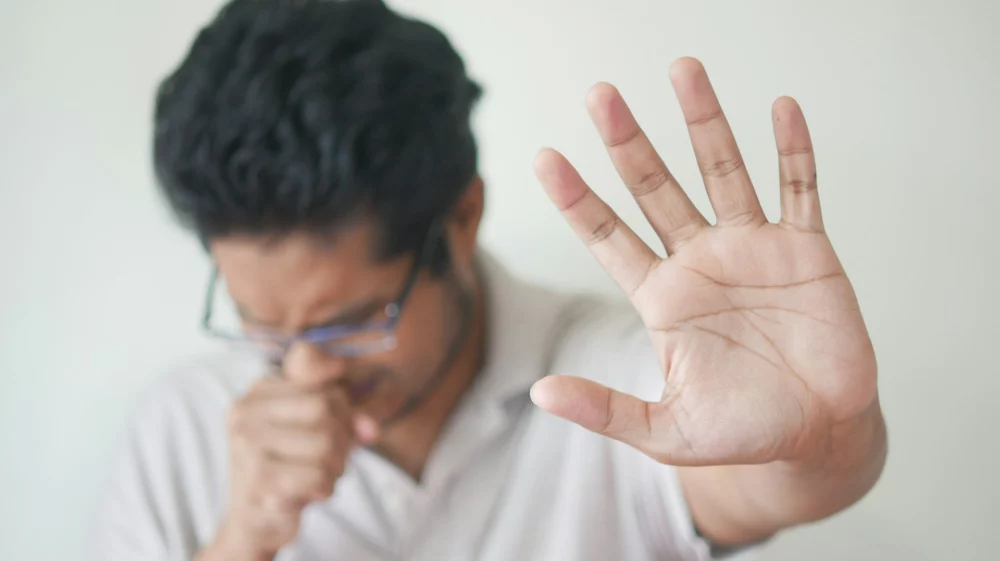 Man coughing into one hand and holding the other hand up towards the viewer.