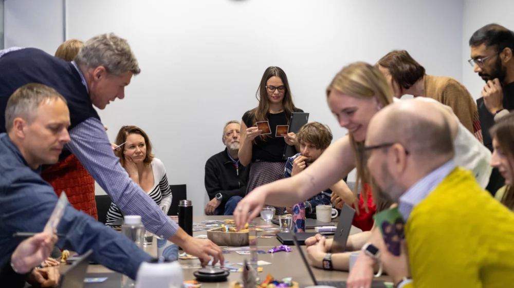 A group of people gathered around a table, collaborating with cards and laptops during a workshop-style meeting.