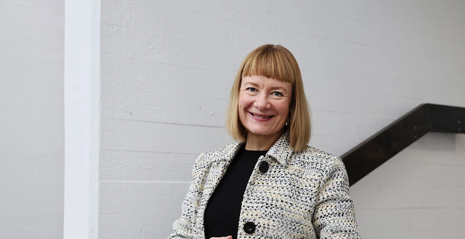 A smiling person with short blonde hair wearing a textured jacket, standing by an indoor staircase.