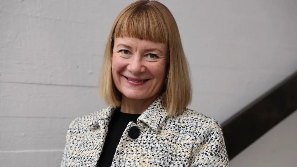 Eeva Primmer smiling with short blonde hair wearing a textured jacket, standing by an indoor staircase.