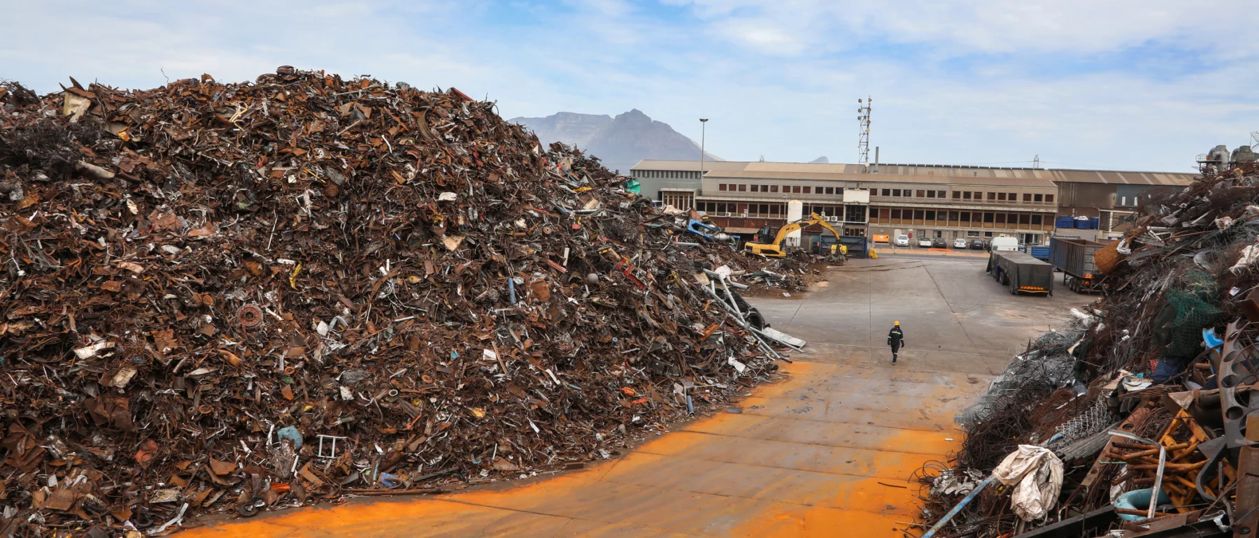 Large piles of rusted scrap metal sit in an industrial recycling yard, with a worker walking between them and a warehouse and mountains visible in the background.