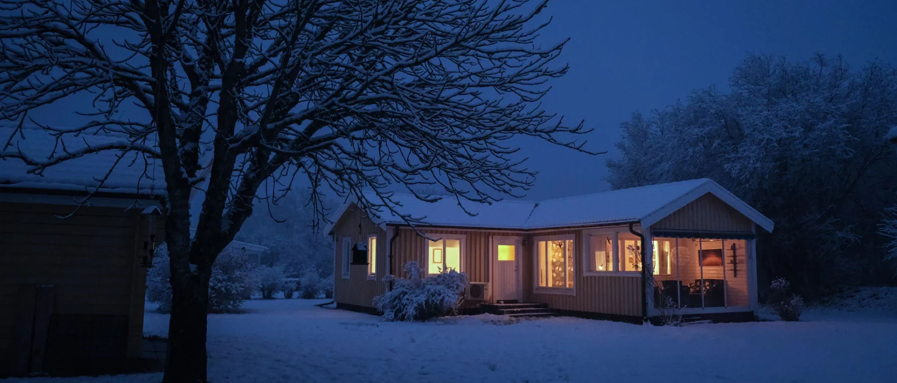 White snow looks almost blue in twilight, on the ground, a tree and the roof of a small yellow house with glowing yellow lit windows.