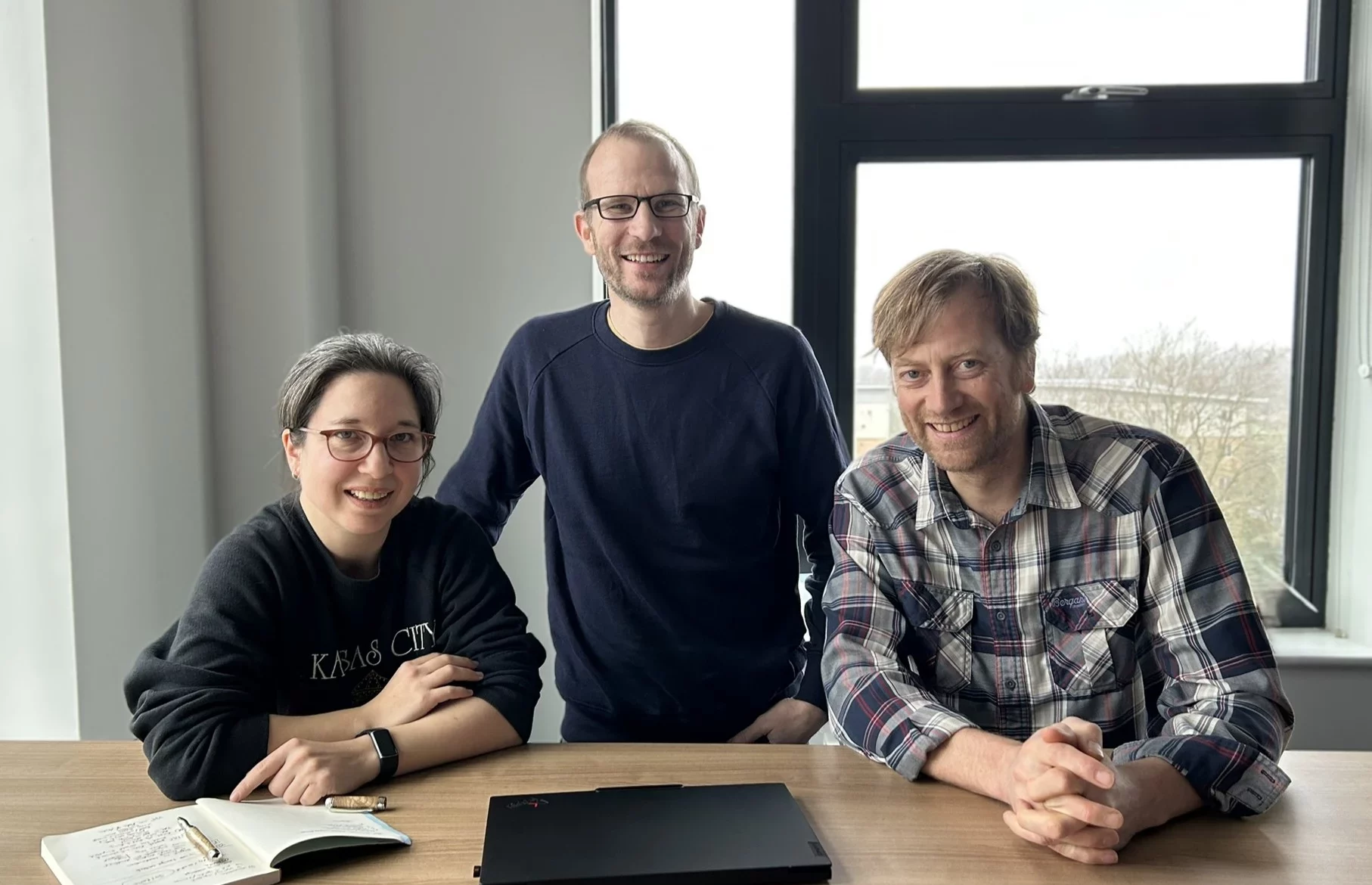 Helen Bellfield, Chris West and Toby Gardner sit at a table indoors, smiling at the camera, with a laptop and notebook on the table and a large window behind them.