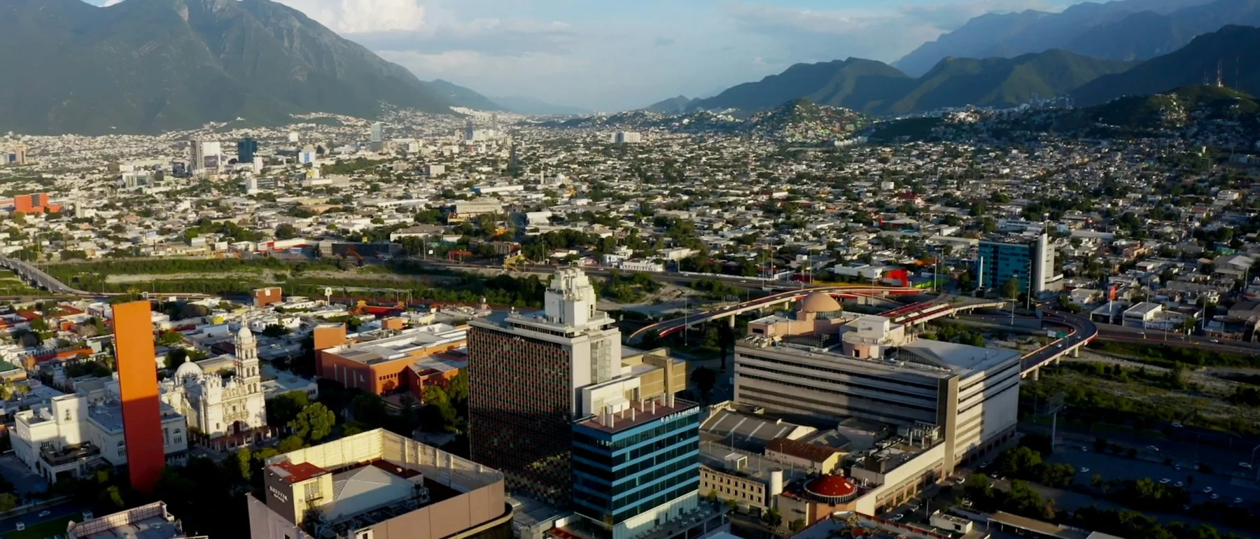 Aerial view of Monterrey, Mexico, featuring a wide sprawl of buildings in the foreground and mountains on the horizon under a lightly clouded blue sky