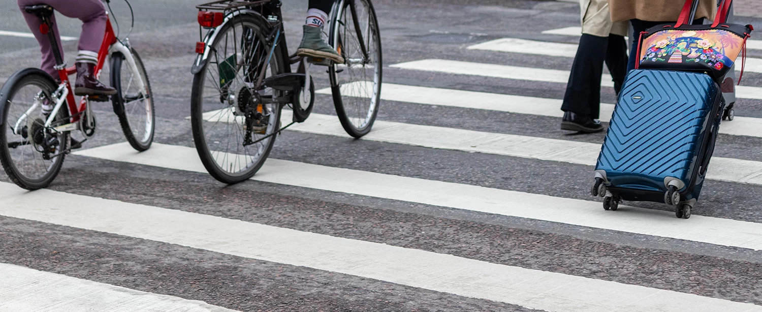 Two cyclists and a person pulling a suitcase crossing a street.