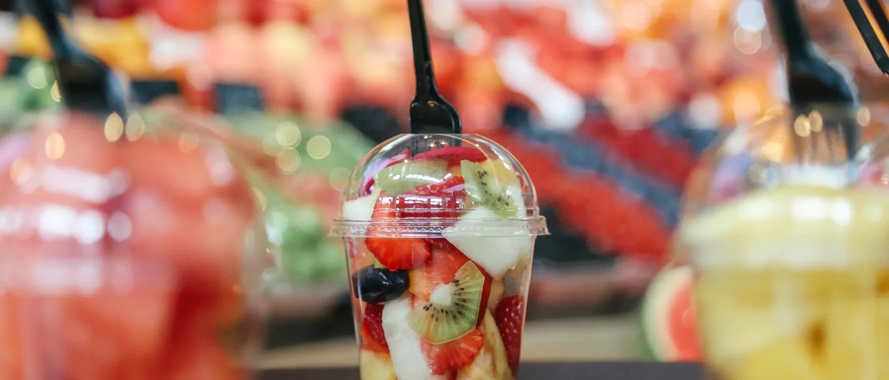 Colorful fruit salad in a plastic see-through cup with a plastic lid, open from the centre. A plastic fork has been stuck in fruit. Around the cup are other cups of fruit and behind are colorful stalls of fruit.