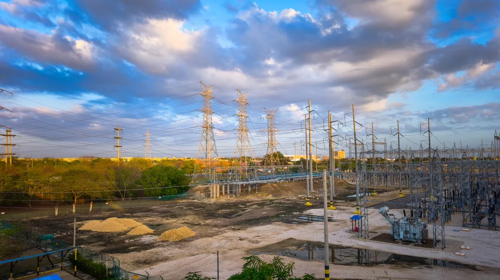 Under a blue sky with clouds, a dozen electricity towers stand above a facility with low buildings and cement aprons, surrounded by green trees.