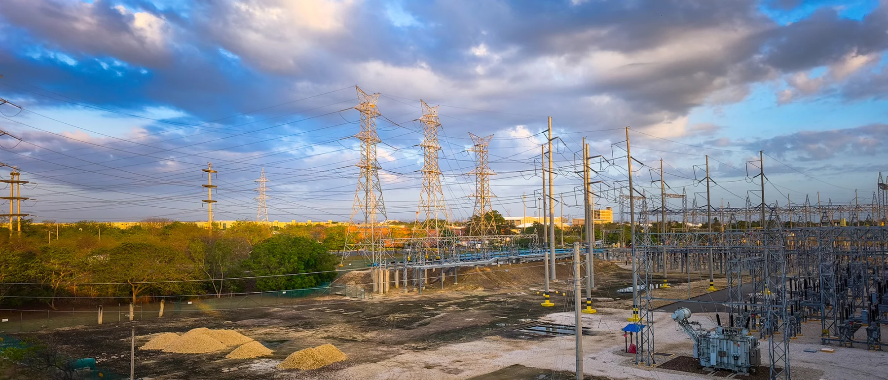 Under a blue sky with clouds, a dozen electricity towers stand above a facility with low buildings and cement aprons, surrounded by green trees.