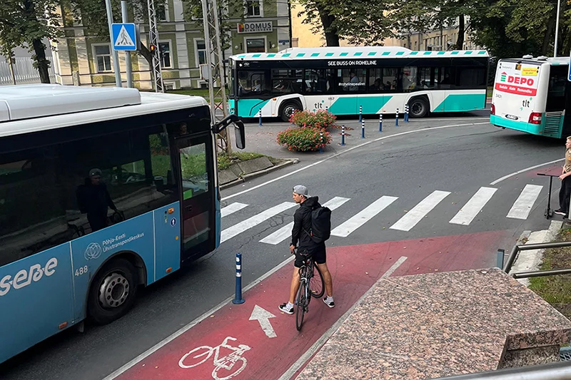 A cyclist is standing at the end of a red cycling path, with an arrow pointing towards a crosswalk. The cyclist is waiting to cross, with a bus blocking the way.