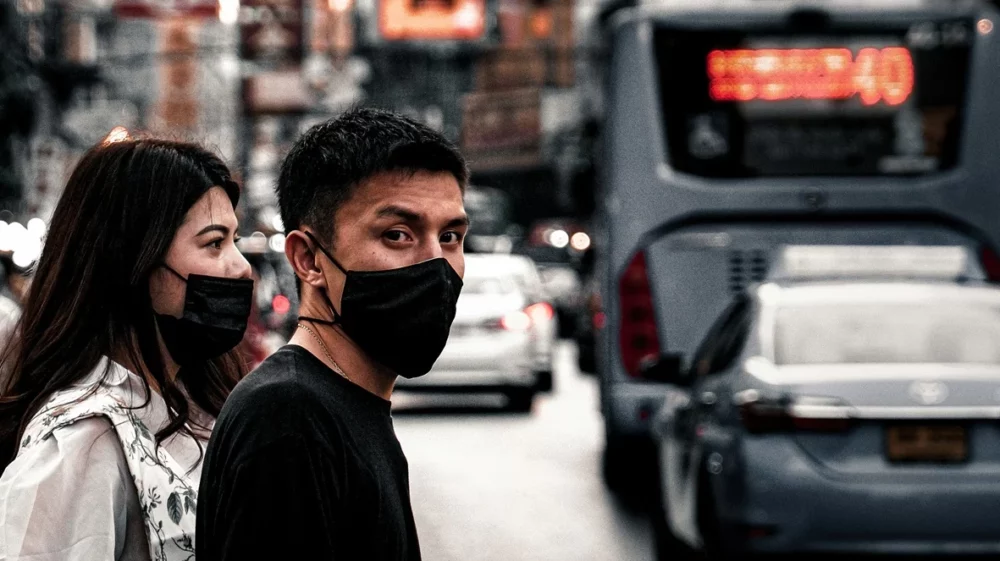 A man in black shirt with a woman in a stone wash grey jacket both wearing mask on a busy street.