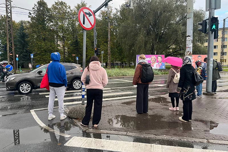 Seven people are waiting on a narrow refugee island at the Kristiine intersection. There are puddles on the traffic island, and the people have spread out around them. One person is standing off the island on the roadway because there is little space and many puddles.