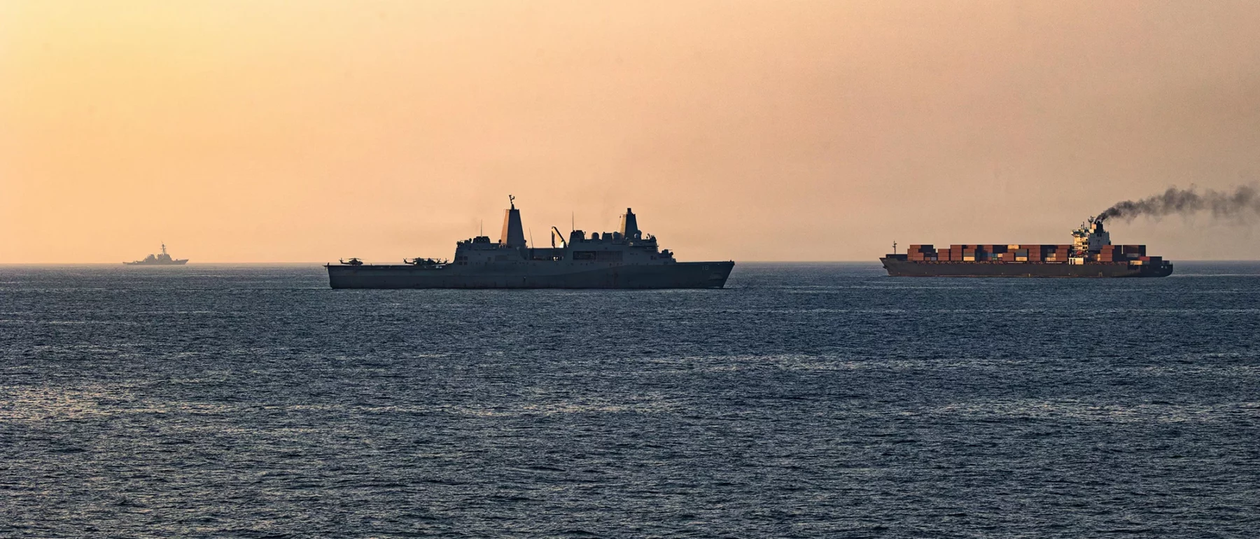 Naval vessels escort a cargo ship at sea during sunset.